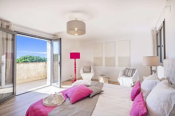 A bright, modern bedroom featuring vibrant pink accents, a bed with a breakfast tray, and open doors leading to a sunny balcony with green trees.