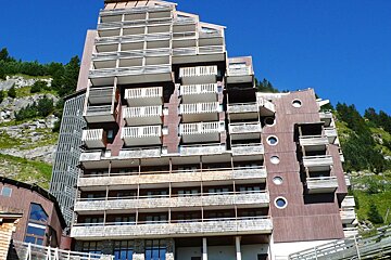 A white multi-story building with wooden balconies by a road, flanked by green/red trees. Mountains and a blue, cloudy sky in the background.
