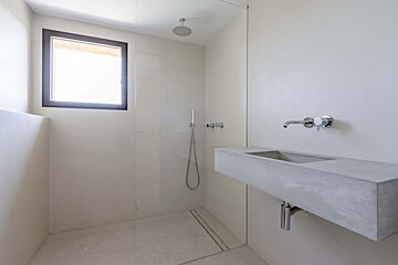 A minimalist modern bathroom with white walls, a window, a glass shower enclosure, and a floating concrete vanity with a wall-mounted faucet.