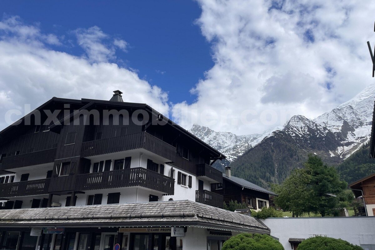 An alpine building with dark wooden facades and white sections, featuring balconies, stands against dramatic snow-capped mountains and a cloudy blue sky.