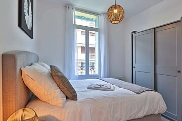 A bright, modern bedroom features a bed with a gray headboard, white bedding, a large window with sheer curtains, a wicker pendant light, and a gray sliding door closet.