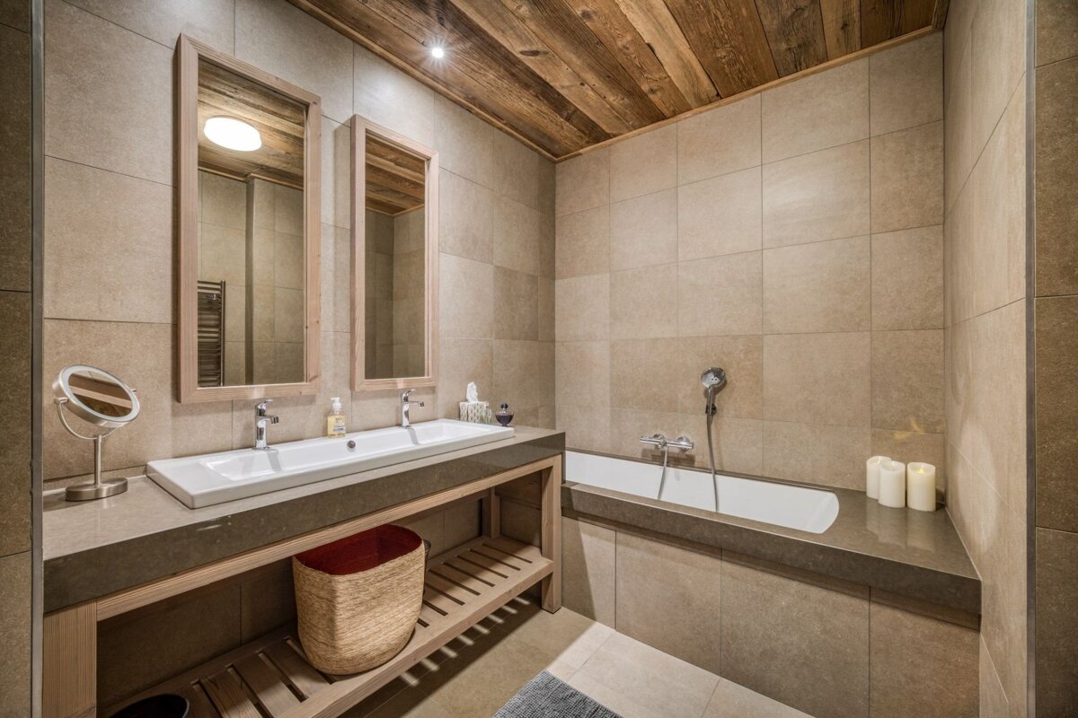 Modern bathroom with a double vanity, a bathtub, neutral tiled walls, and a rustic wooden ceiling, offering a warm, cozy ambiance.