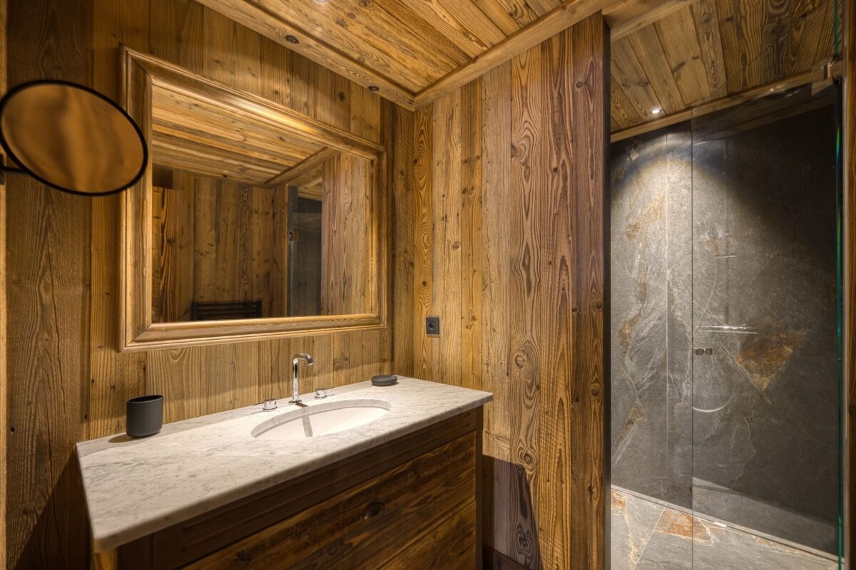 Rustic chalet-style bathroom featuring wood-paneled walls, a marble-top vanity with a large mirror, and a dark stone-tiled glass shower.