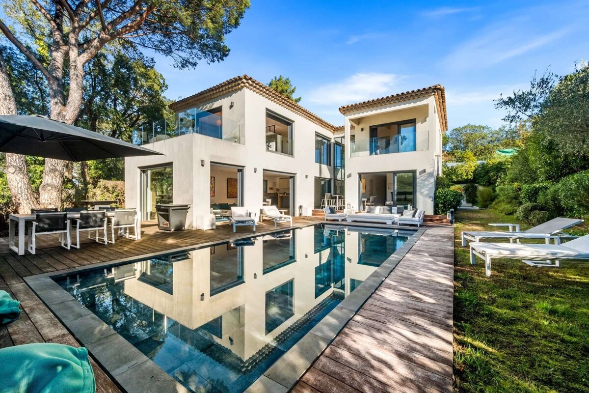 A modern white house with a swimming pool, wooden deck, and outdoor seating under a sunny sky. Lush greenery surrounds the property.