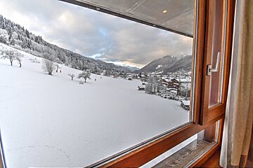 A snowy winter landscape with mountains and a distant village, seen through a wooden window.