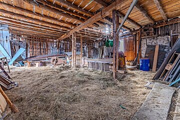 A messy, rustic barn interior with hay on the floor, exposed wooden beams, scattered tools, a bike, and a blue barrel.