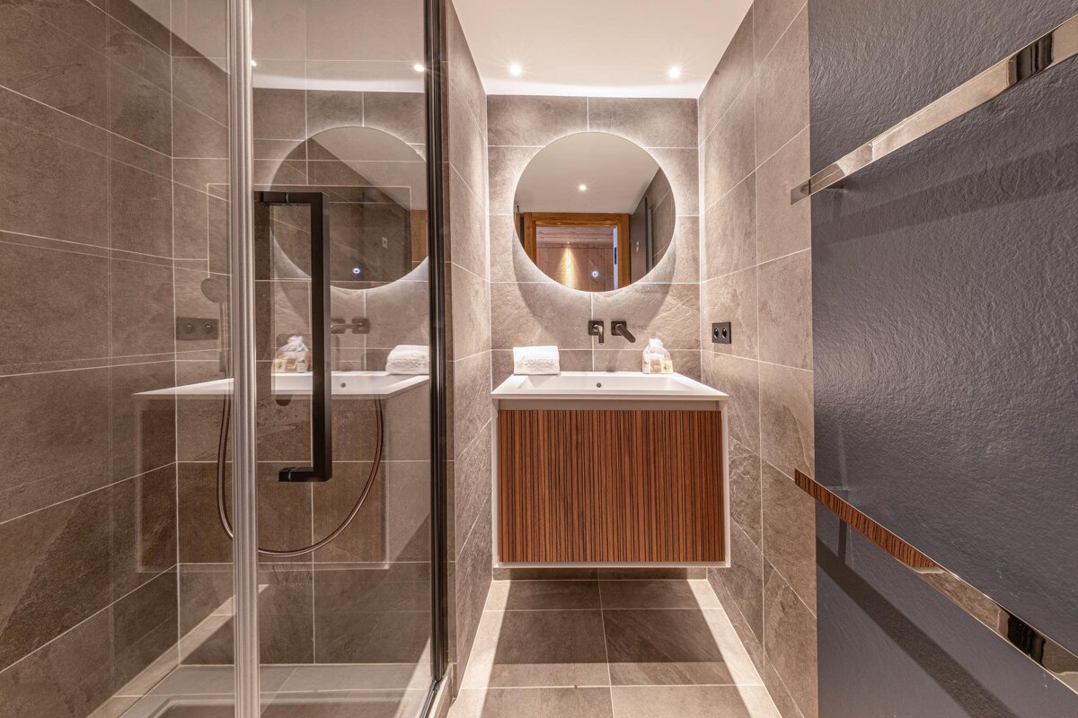 Modern tiled bathroom with a glass shower, floating wooden vanity, and a backlit circular mirror.