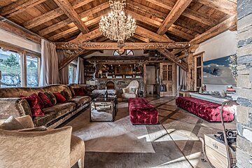 Luxurious chalet living room with exposed wooden beams, a grand chandelier, velvet sofas, and red ottomans, creating a rustic-chic ambiance.