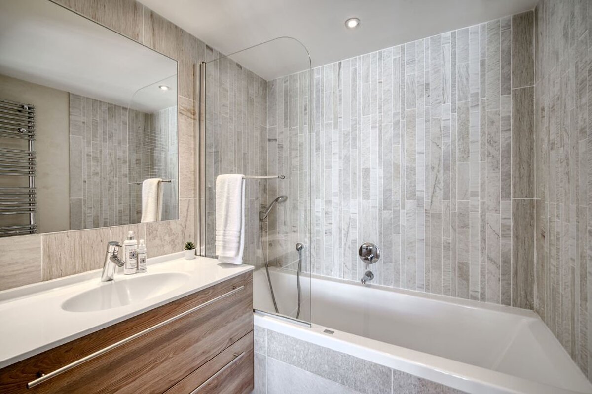 Modern bathroom with wood vanity, white sink, large mirror, and a bathtub with glass shower screen. Walls are covered in light grey tiles.