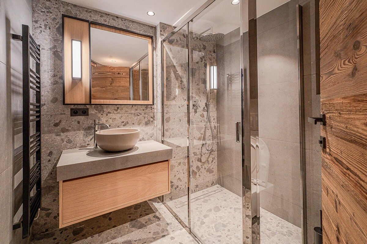 Modern bathroom with terrazzo-style grey stone tiles, a wooden vanity with bowl sink, a mirror, and a glass-enclosed walk-in shower.