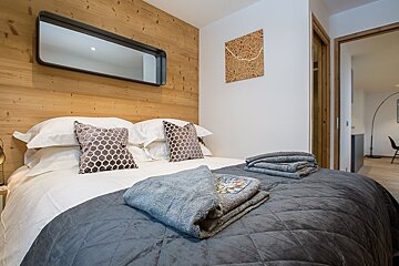 Modern bedroom featuring a wood-paneled accent wall with a mirror, a double bed with grey throws, towels, and patterned pillows.