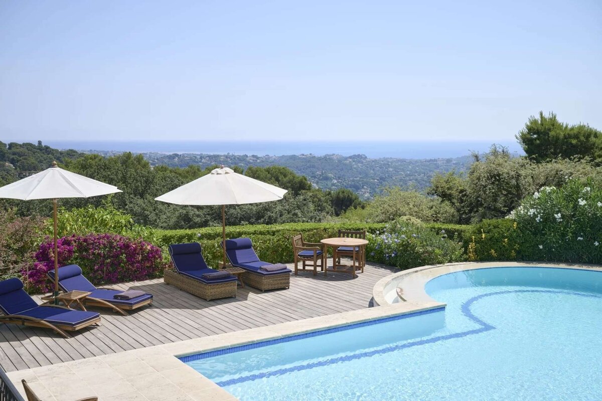 Sunny outdoor pool area with a wooden deck, lounge chairs, umbrellas, and dining set, overlooking a lush landscape and the distant sea.