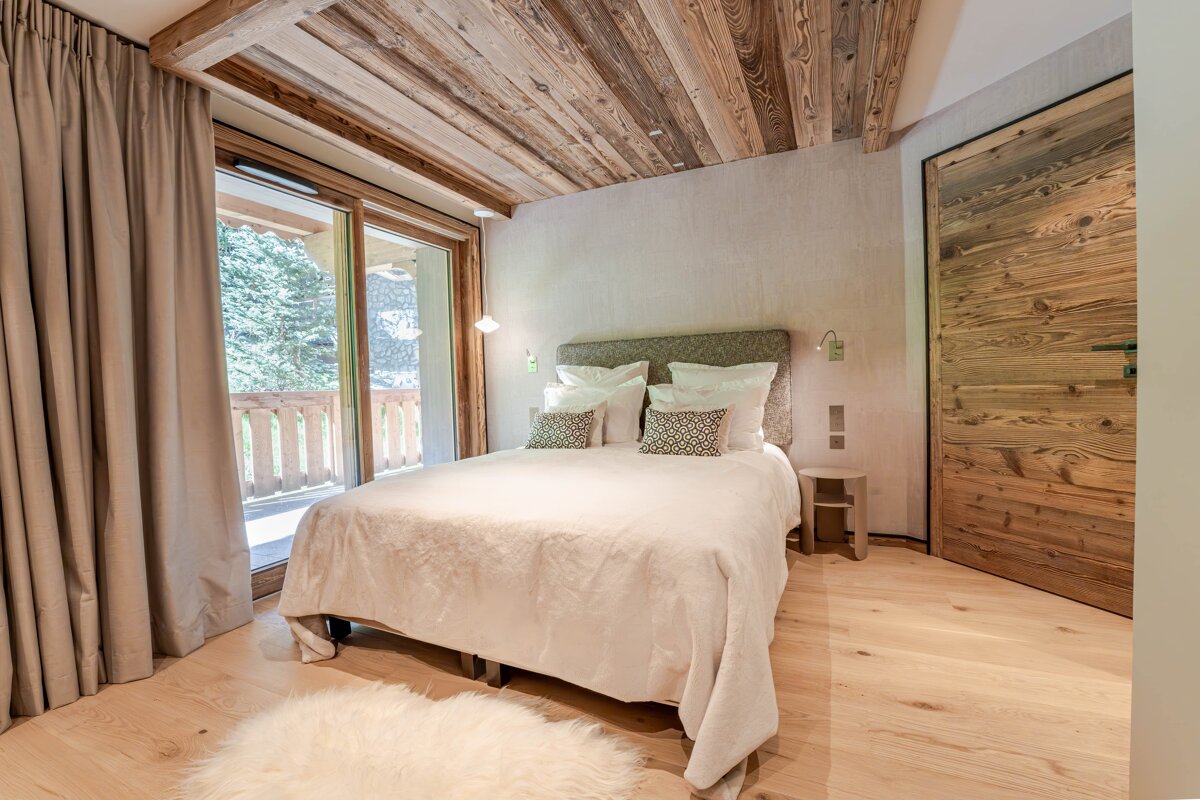 A warm, modern chalet bedroom with a plush bed, rustic wooden ceiling/floor/door, and a balcony with tree views. A white shag rug accents the floor.