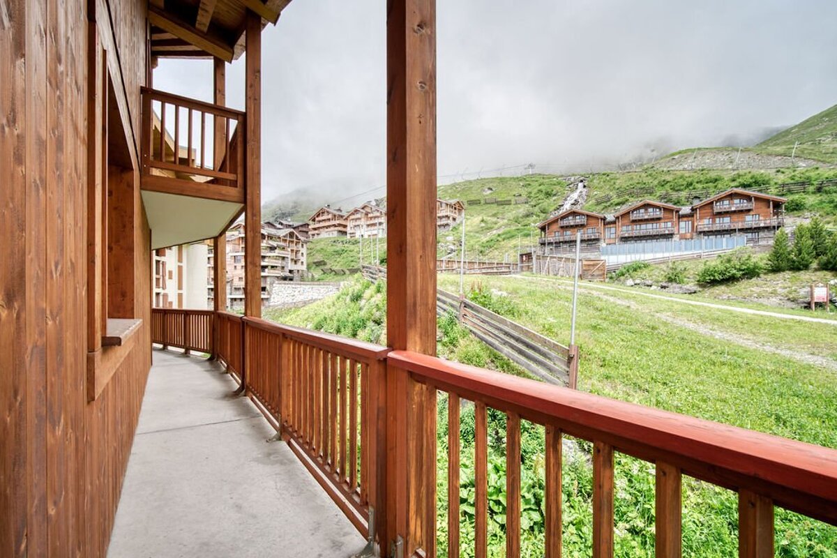View from a wooden balcony overlooking chalet buildings nestled on green mountain slopes under a cloudy sky, with mist hugging the peaks.