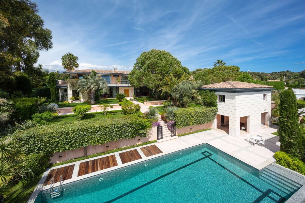 An aerial view of a luxurious villa with extensive, manicured gardens and a large, sparkling blue swimming pool under a clear sky.