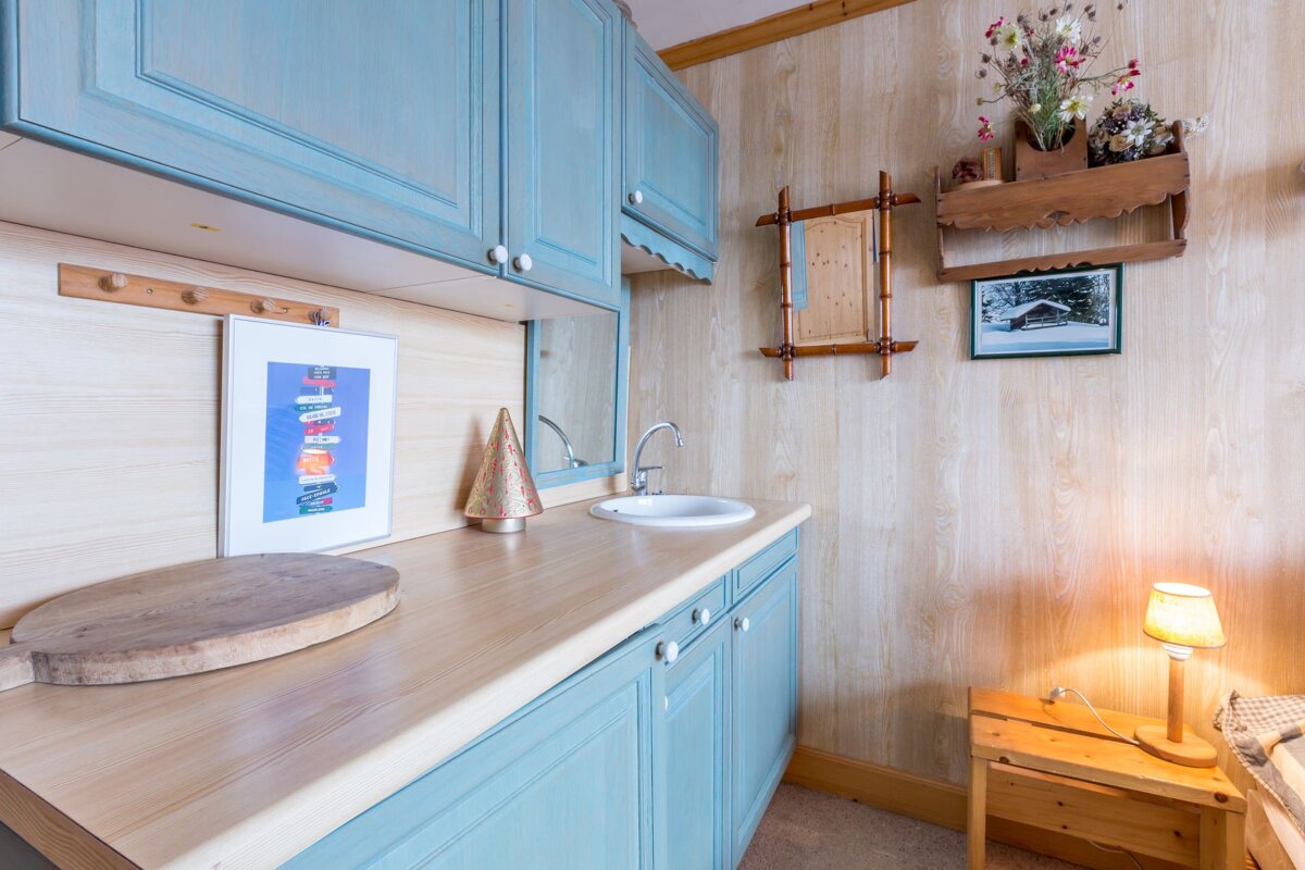 A rustic kitchenette features light blue cabinets, wooden walls/counter, a sink, and charming decor including framed art, flowers, and a bedside lamp.