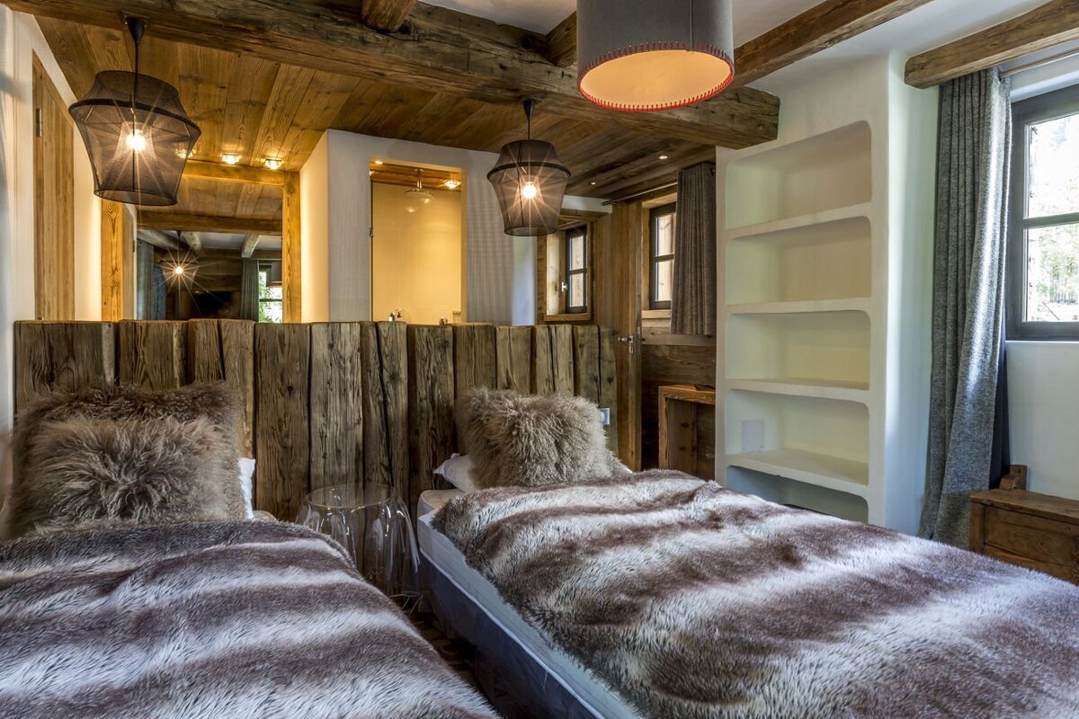 Cozy chalet bedroom with twin beds, plush faux fur bedding, rustic wooden walls/ceiling, and modern pendant lights. Built-in shelves visible.