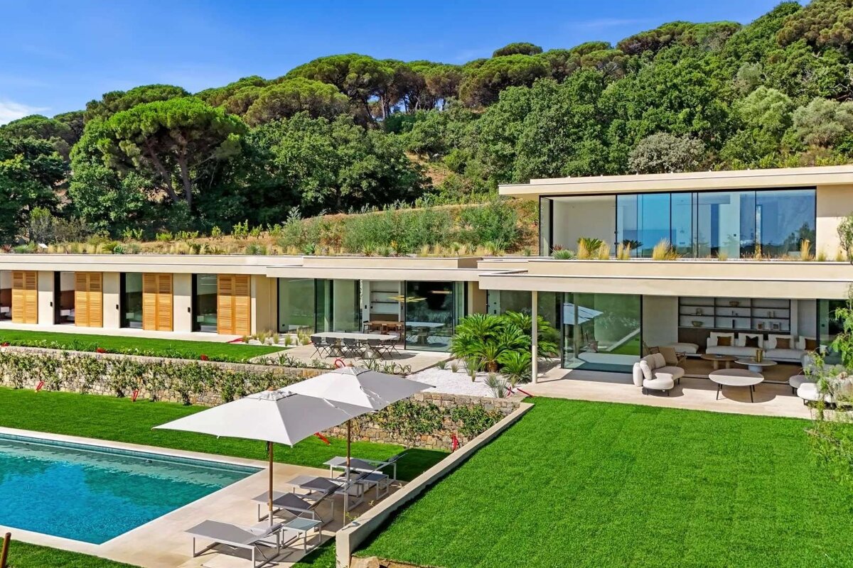 A modern, multi-level villa with green roofs, large windows, a swimming pool, and manicured lawns, set against a lush, forested hillside.