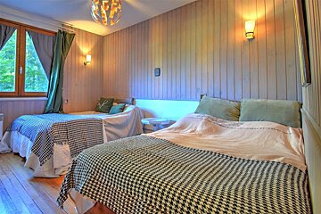Cozy bedroom featuring two beds with houndstooth blankets, wood-paneled walls, warm lighting, and a window with green curtains.