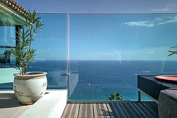 Modern balcony with glass railing showcasing a vast blue ocean and sky. A potted plant and wooden deck are bathed in sunlight.