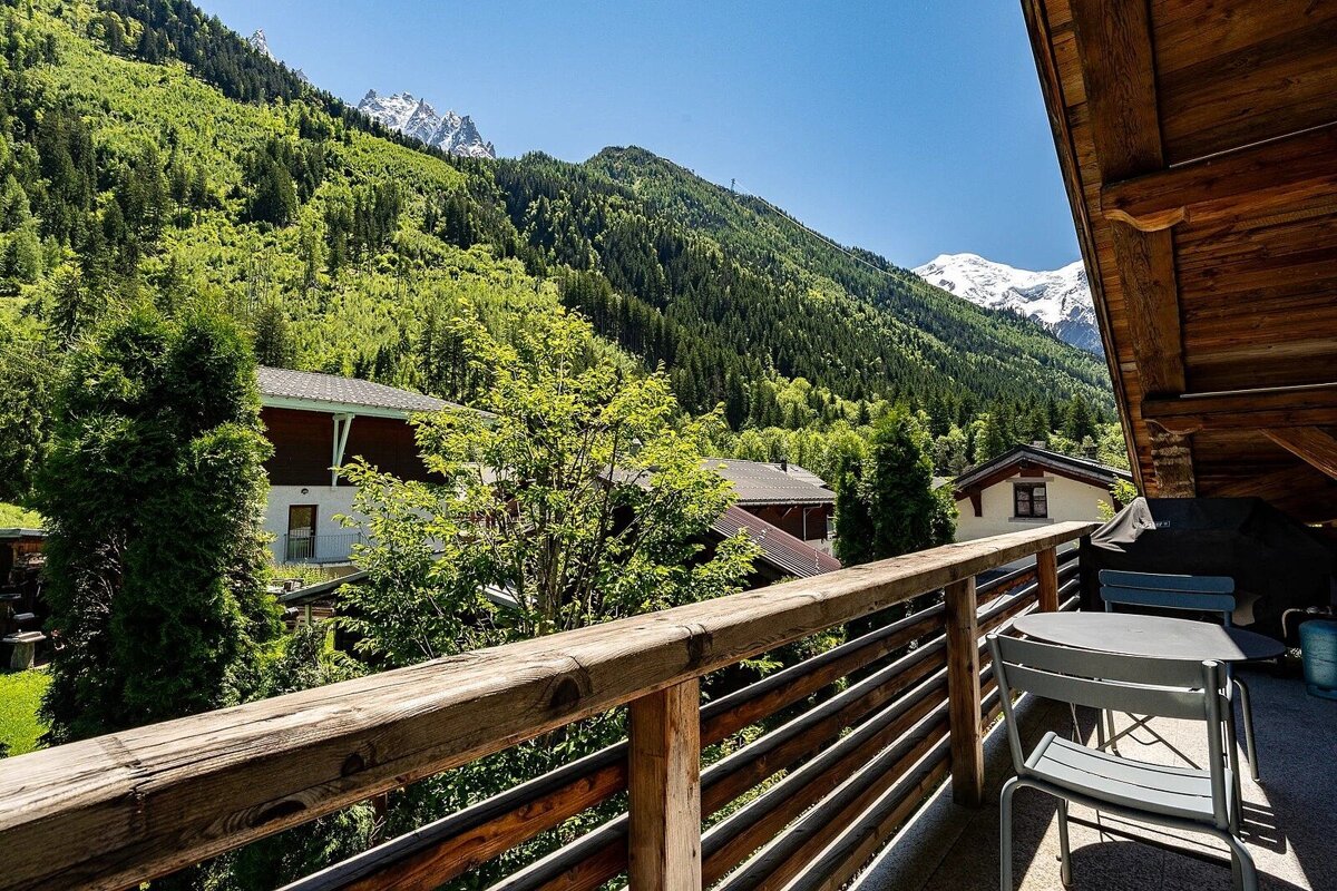 A sunny balcony overlooks a green mountain valley with houses and distant snow-capped peaks under a clear blue sky.