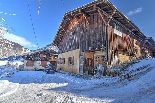 A rustic wooden barn and house are nestled in a snowy mountain landscape under a bright blue sky, with a car parked on the snow.