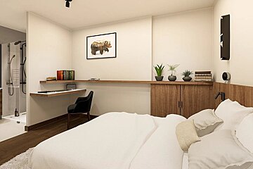 A modern, minimalist bedroom featuring a white bed, integrated desk, plant decor, and bear artwork, with an en-suite shower visible on the left.
