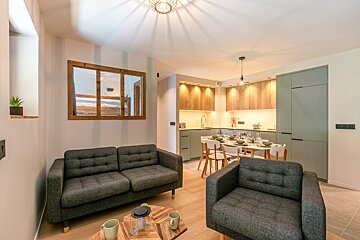 Modern open-plan apartment featuring a dark grey sofa and armchair, a dining area, and a kitchen with sage green and wood cabinets.