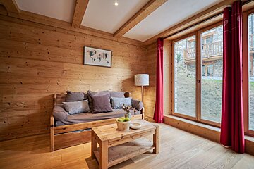 Cozy, wood-paneled room with a sofa, wooden coffee table holding apples, and a large window with red curtains looking out to a stone building.