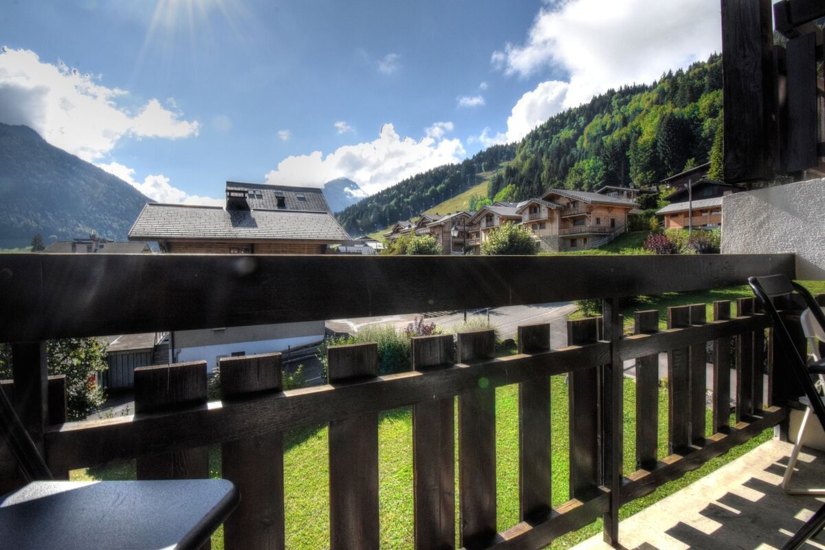 View from a wooden balcony of alpine chalets and lush green mountains under a bright sunny sky.