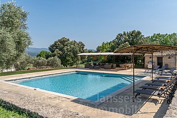 A beautiful blue swimming pool area with stone paving, outdoor lounge and dining, sun loungers, and a stone pool house, surrounded by lush trees under a clear sky.
