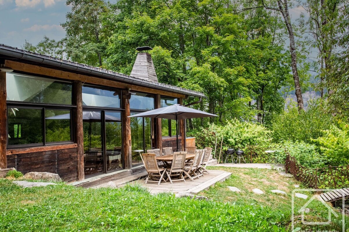 A rustic wooden cabin with large windows, an outdoor dining deck under an umbrella, surrounded by lush green trees and grass.
