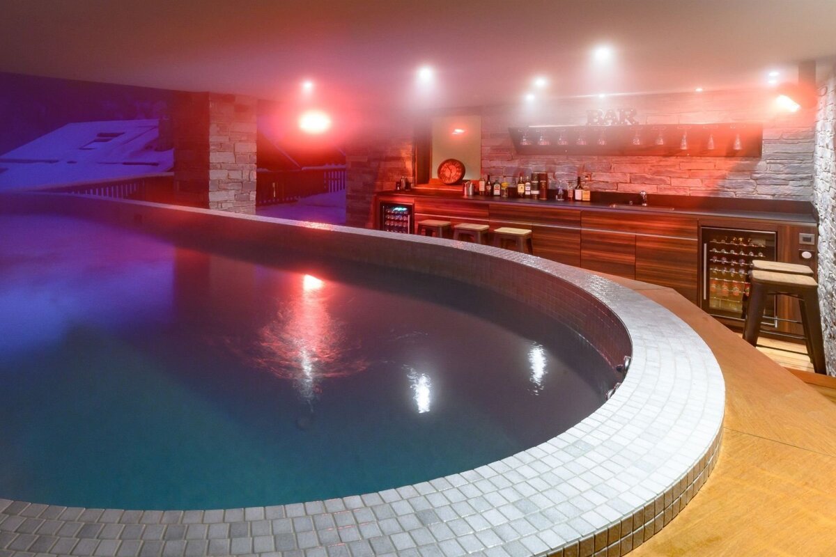 Luxurious indoor pool with red and blue lighting, alongside a well-stocked bar, and a view of a snowy outdoor scene.