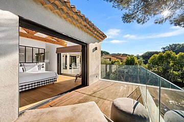 Sunlit bedroom opens to a balcony with glass railing, offering scenic views of green trees and distant houses under a clear blue sky.