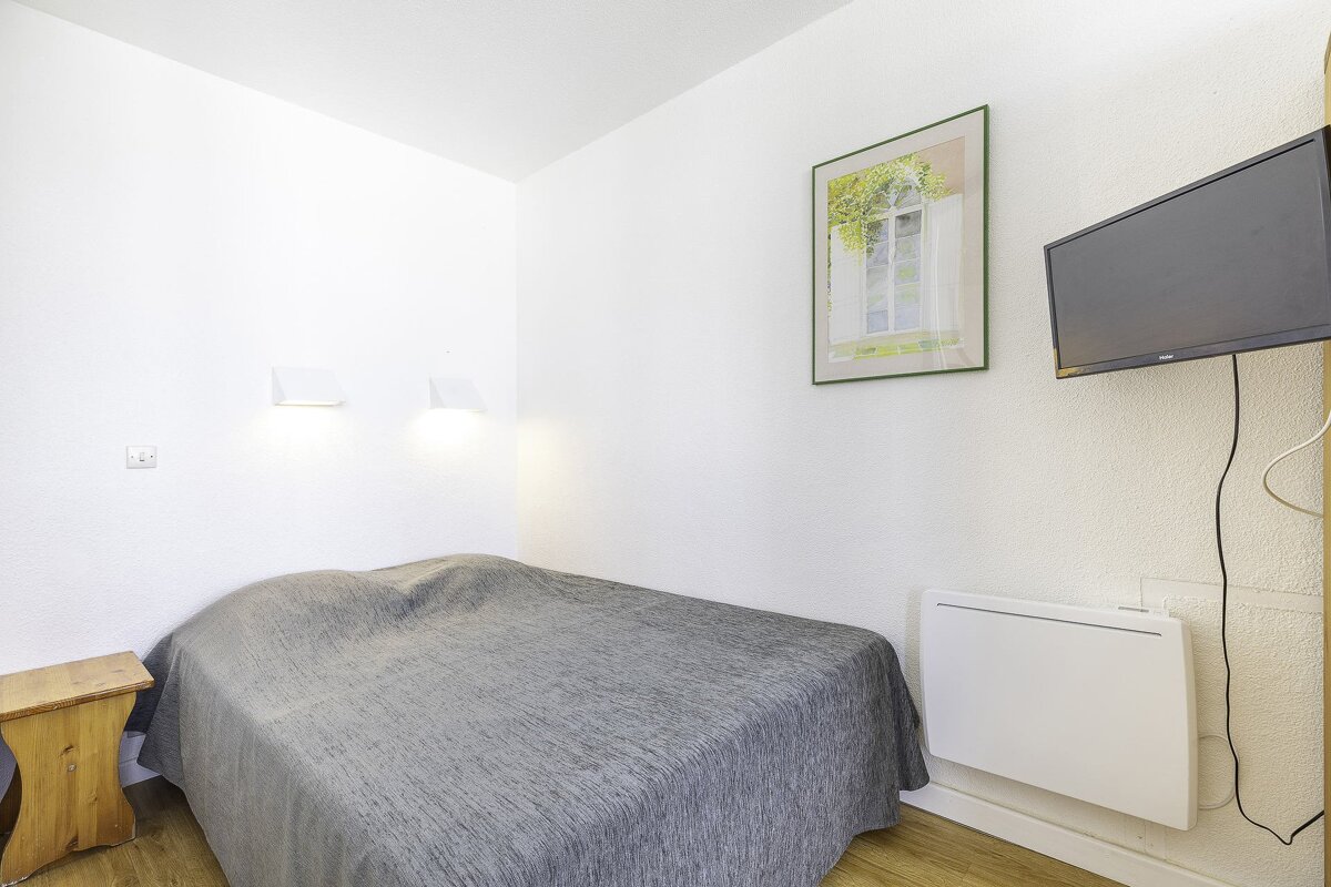 A small, minimalist bedroom with white walls, a grey bed, wooden side table, wall-mounted TV, heater, and a framed picture.