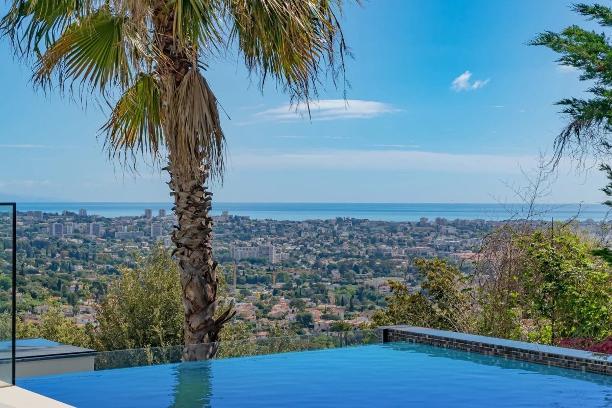 An infinity pool overlooks a coastal city and the ocean under a blue sky, framed by a palm tree in the foreground.