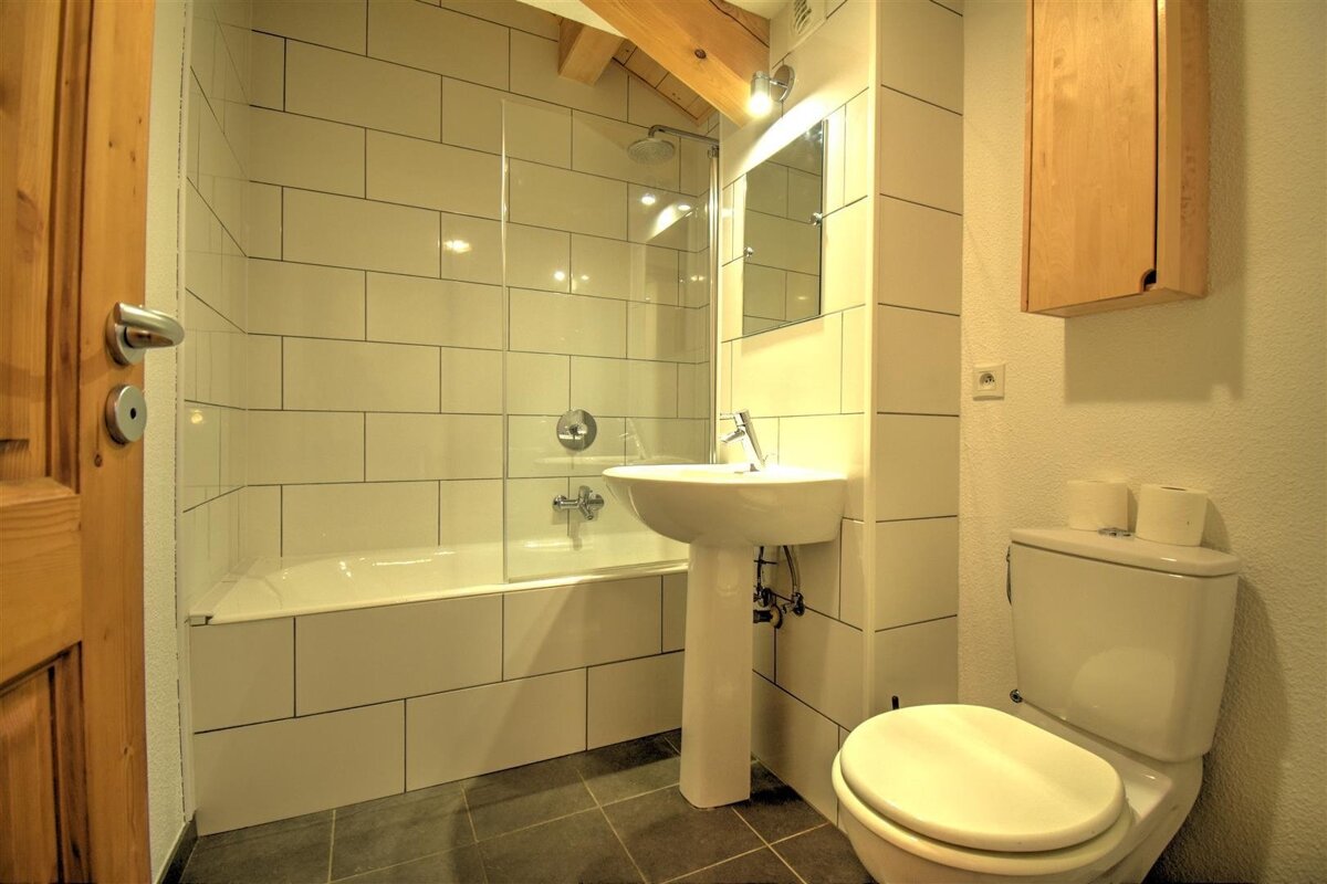 A bright, modern bathroom featuring white subway tiles, a bathtub with glass partition, pedestal sink, toilet, and a wooden door.