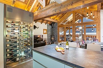 Modern rustic chalet kitchen featuring a large glass-front wine cellar, stone island, and exposed wooden beams.