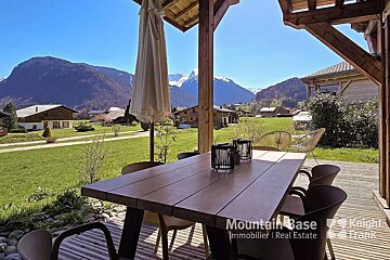 A sunny mountain view from a covered patio with a wooden table. Green fields, distant houses, and snow-capped peaks fill the landscape under a clear blue sky.