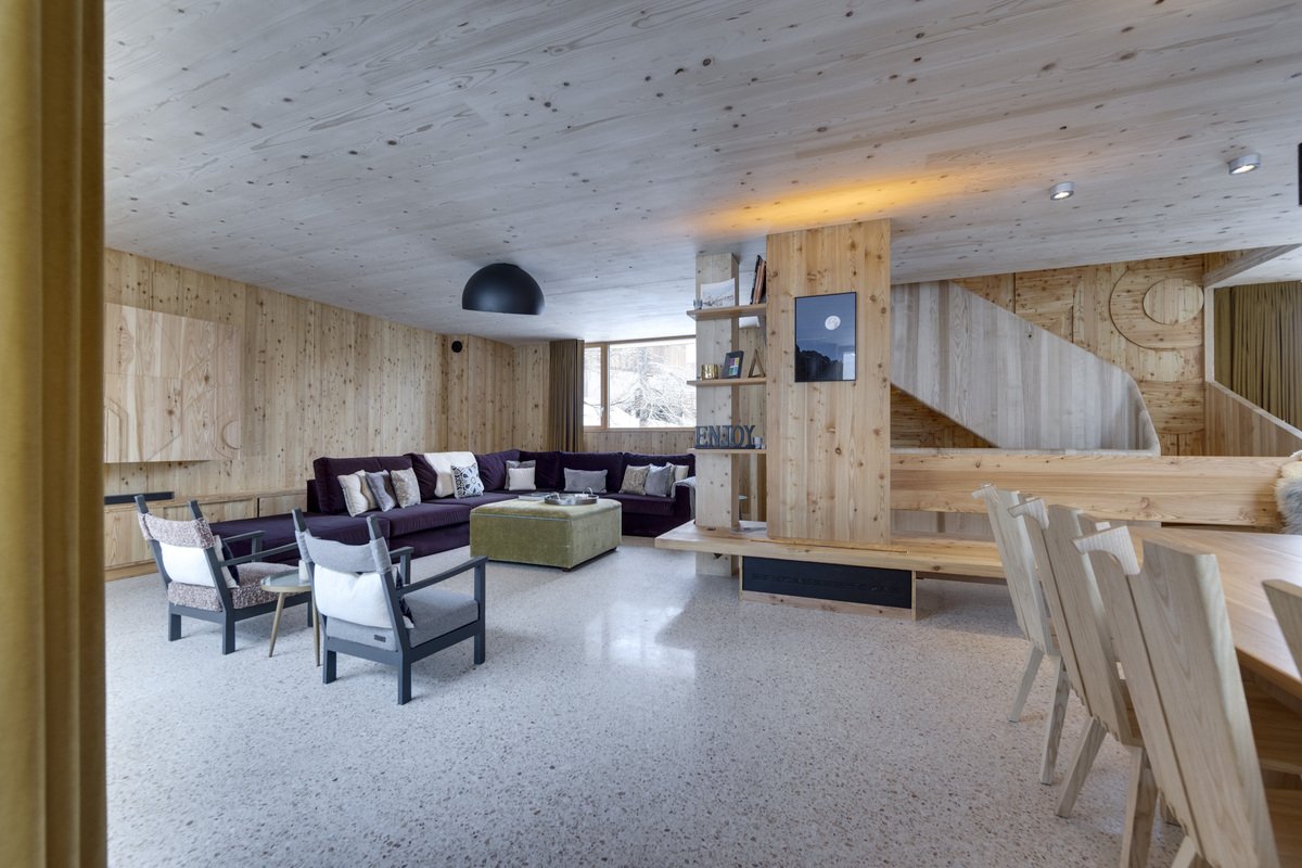 Modern chalet interior with light wood-paneled walls and ceiling, a large dark sofa, terrazzo flooring, and wooden dining chairs.