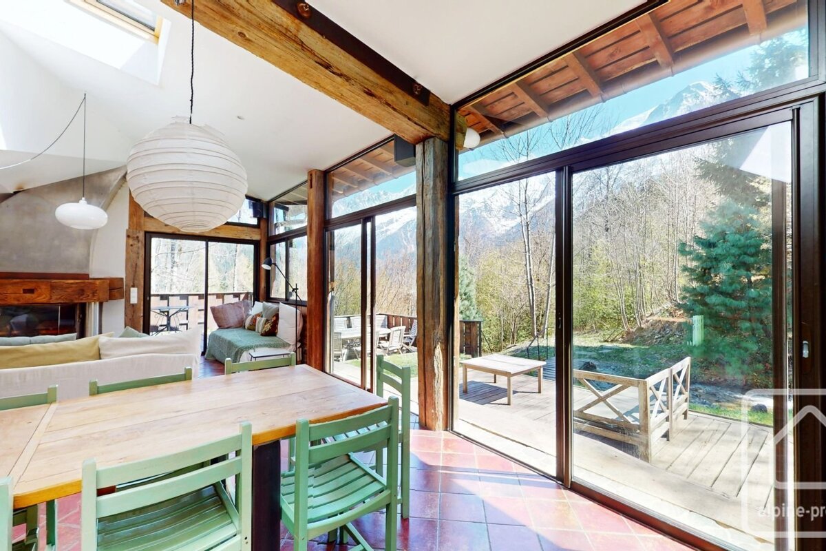 Rustic dining room with vast windows showcasing snowy mountains and a forest. Wooden beams, light green chairs, and an outdoor deck.