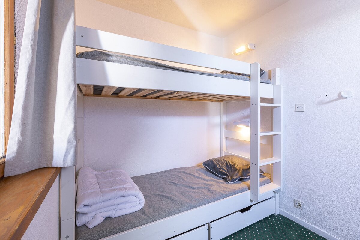 A simple room with white wooden bunk beds. The bottom bunk has gray bedding, a folded white duvet, and a pillow. A ladder, wall lights, and green carpet are visible.
