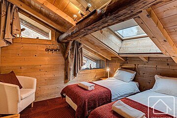 A cozy, rustic wooden bedroom with slanted ceilings, two single beds, skylights, and exposed beams, offering a warm chalet ambiance.