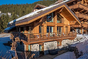 A beautiful wooden and stone chalet with a snowy roof and balcony, nestled among evergreen trees on a clear, sunny winter day.