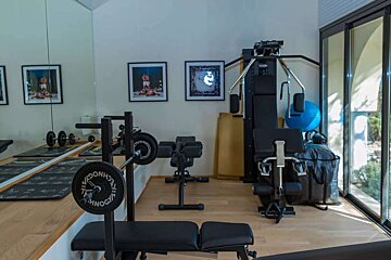 A modern home gym with a barbell, bench, multi-gym, and stability ball, reflecting in wall mirrors and illuminated by large windows.