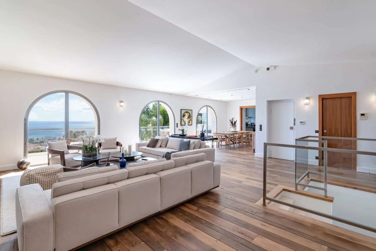A bright, modern open-plan living room with arched windows offering ocean views. Features light sofas, wooden floors, a dining area, and a staircase.
