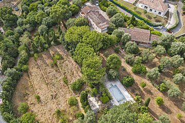 Aerial view: Houses with red roofs, an empty pool, and dry terraced fields among lush green trees, including olive groves.