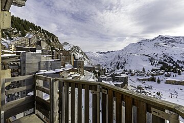 View of a snowy mountain ski resort and wooden buildings from a balcony under a cloudy sky.