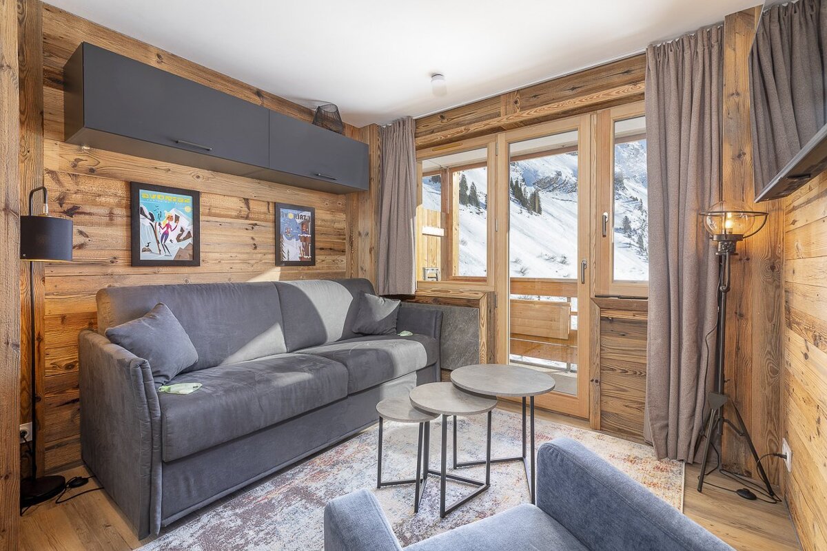 Alpine living room with warm wooden walls, a gray sofa, and a large window showcasing snow-covered mountains.