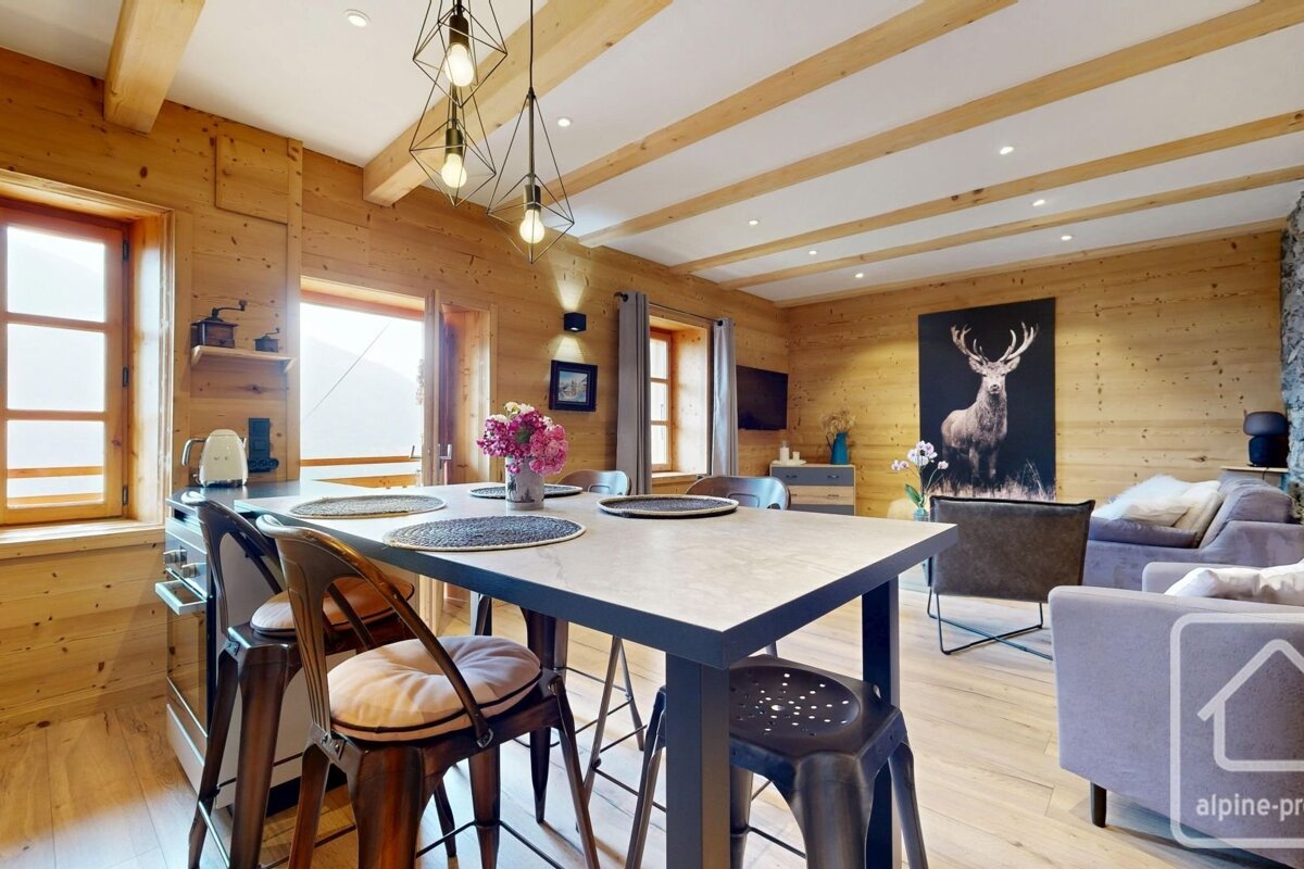 Cozy chalet interior with open dining/living areas, warm wood paneling, deer art, and bright windows.